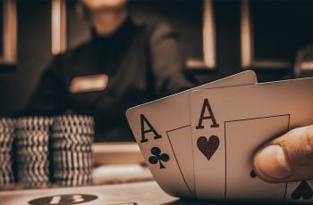 Zwei Asse als Startkarten auf einem Pokertisch, umgeben von bunten Chips, während andere Spieler sich auf ihre Karten konzentrieren.