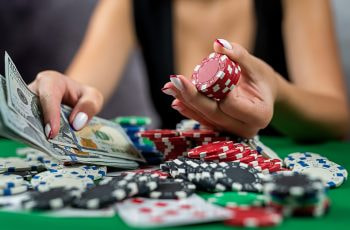 Eine weibliche Person sitzt vor einem Pokertisch mit vielen Pokerchips und Karten und hält eine Menge Geldscheine in der Hand.